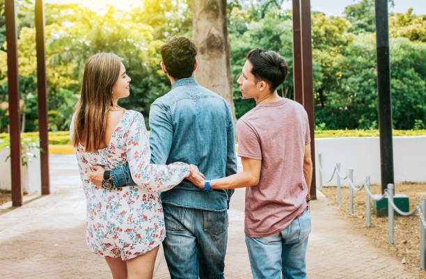 unfaithful girl walking in the park with her boyfriend while holding another man hand. love triangle concept. woman holding hands with another man while walking with her boyfriend outdoor - infidelidade imagens e fotografias de stock