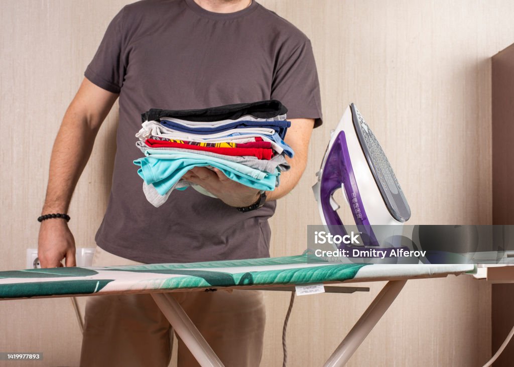 a man holds a stack of ironed laundry against the background of an ironing board and an iron a man holds a stack of ironed laundry against the background of an ironing board and an iron. copy space. Adult Stock Photo