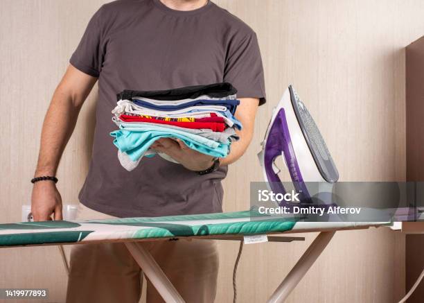 A Man Holds A Stack Of Ironed Laundry Against The Background Of An Ironing Board And An Iron Stock Photo - Download Image Now