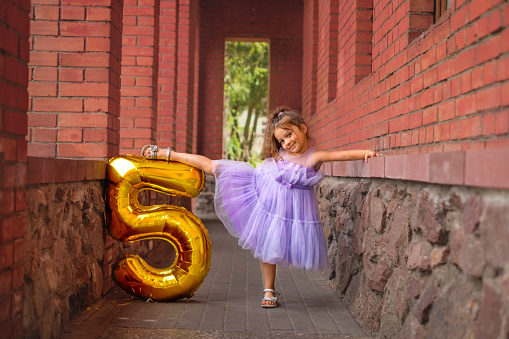 A five-year-old gymnast girl in a purple festive outfit with golden balloon in the shape of number stands in the pose of a ballerina. Child celebrates the first anniversary