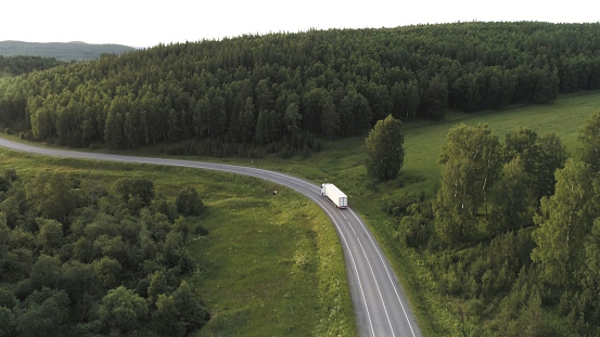 Wagon driving on the highway surrounded by green trees, aerial view. Transport, logistics concept, white truck driving on the empty road along green forest.