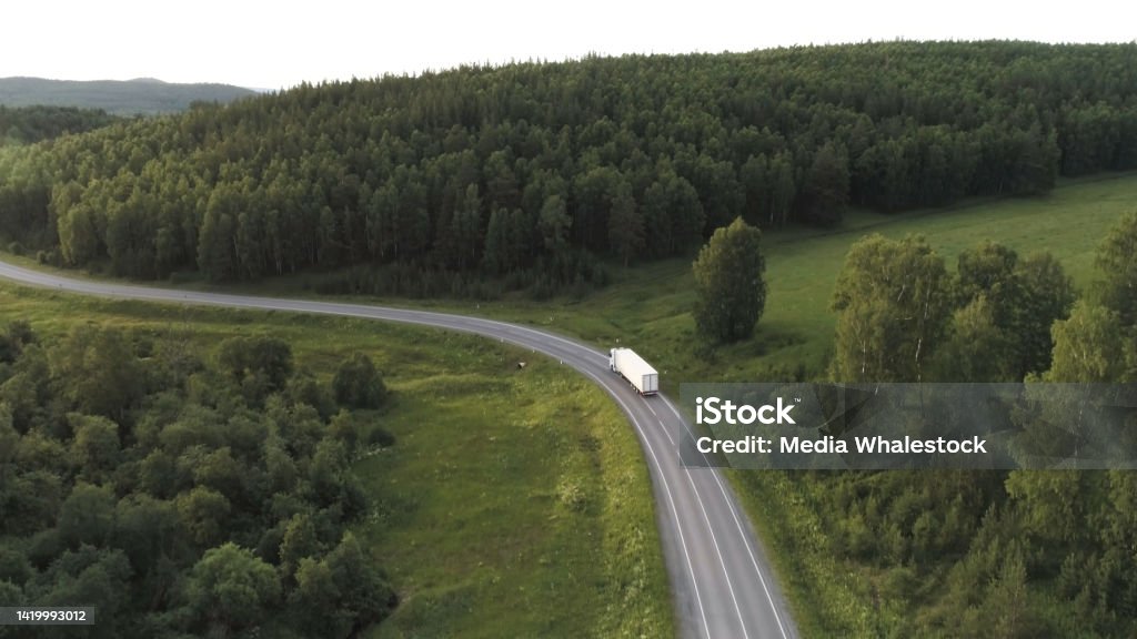 Wagon driving on the highway surrounded by green trees, aerial view. Scene. Transport, logistics concept, white truck driving on the empty road along green forest. Wagon driving on the highway surrounded by green trees, aerial view. Transport, logistics concept, white truck driving on the empty road along green forest. Mountain Peak Stock Photo
