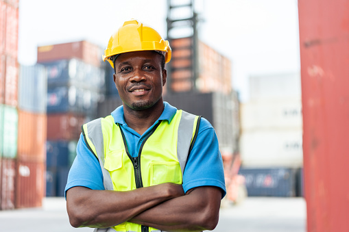 Portrait of Young African American worker work in container termainal. Attractive male laborer processing orders and products to plan at warehouse logistic in cargo freight ship for import export.