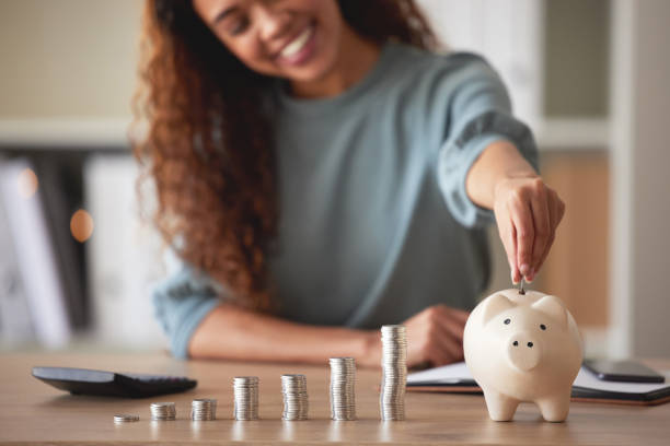 Young african american woman money putting coins into a piggybank at home. Mixed race person counting coins while financial planning in her living room. Saving, investing and thinking about the future Young african american woman money putting coins into a piggybank at home. Mixed race person counting coins while financial planning in her living room. Saving, investing and thinking about the future counting stock pictures, royalty-free photos & images