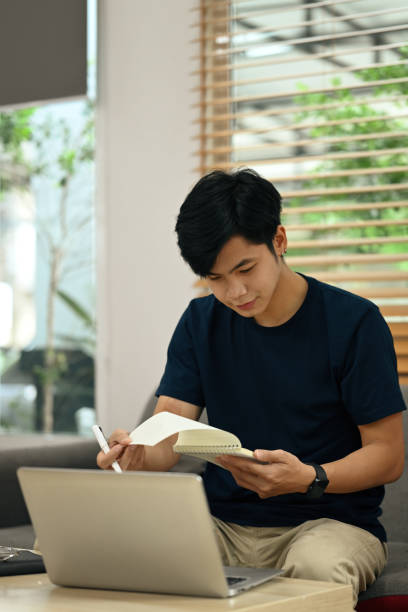 istock Focused asian male freelancer working online on laptop and making notes on notepad. 1420034091