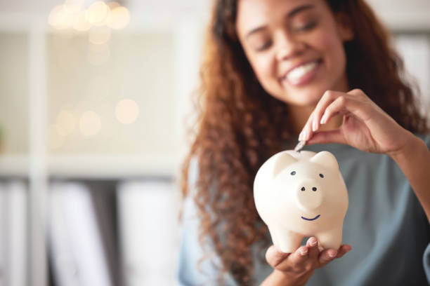 one happy young mixed race woman holding a piggybank and depositing a coin as savings. hispanic woman budgeting her finances and investing money into her future. saving funds for financial freedom - huishoudkosten stockfoto's en -beelden