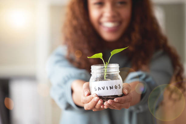 joven afroamericana presentando su frasco de ahorros de vidrio con una planta en ciernes que crece de él en casa. feliz persona de raza mixta sonriendo mientras planifica, ahorra e invierte para su futuro - twenty first fotografías e imágenes de stock