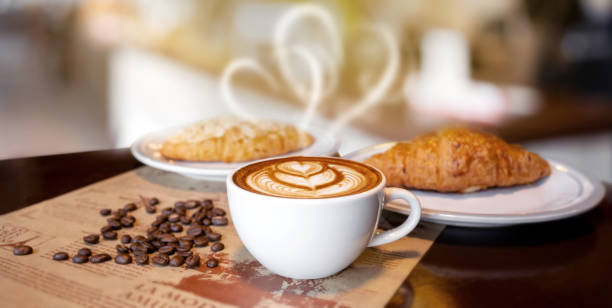 Hot latte coffee in white cup with heart shape smoke, with bakery and coffee beans placed on dark brown wood table, bakery and natural food, with light and dark,Italian and french cafe and croissant.