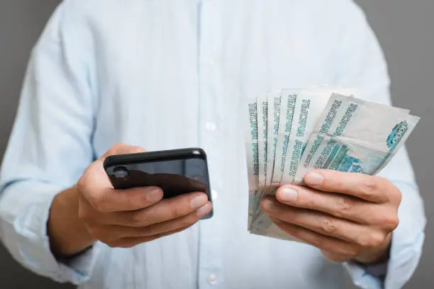 Photo of Business, finance and technology concept. Close-up of man holding thousands of rubles and using smartphone while standing indoors on gray background. Selective focus on money and mobile phone