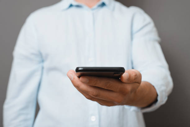 istock Unrecognizable man in blue shirt holding smartphone while standing against gray background indoors, close-up. Front view businessman showing mobile phone. Selective focus on device 1420053345