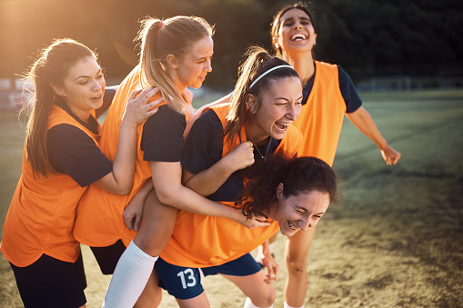 Team of cheerful female players celebrating scoring a goal during soccer match on playing field.