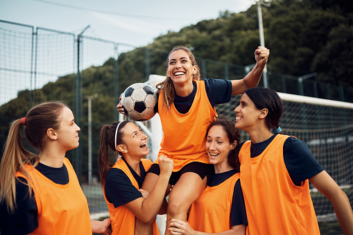 Happy women's soccer team carrying one of the players while celebrating wining the match on playing field.