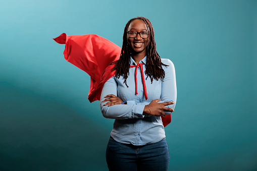 Confident looking mighty powerful young adult person wearing superhero costume on blue background. Portrait of happy brave woman with superpowers wearing red hero cape while standing with arms crossed.