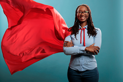 Brave and happy looking young adult superhero woman wearing red hero cape while smiling at camera. Justice defender with superpowers and mighty posture standing with arms crossed on blue background.