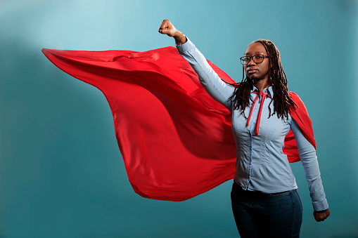 Strong justice defender with superpowers looking at camera with fist in air. Brave and proud looking young adult superhero woman acting like a flying hero while wearing mighty cape on blue background.