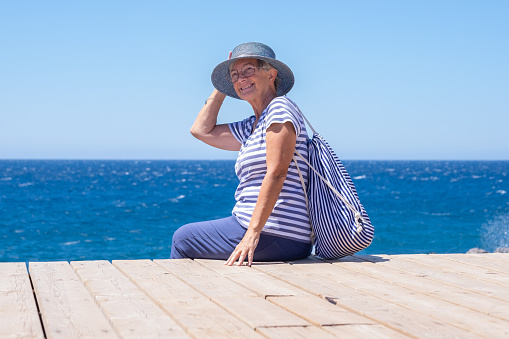 Happy senior woman dressed in blue sitting at the sea holding her hat lest it fly away. Mature lady with eyeglasses enjoying freedom and relax in vacation