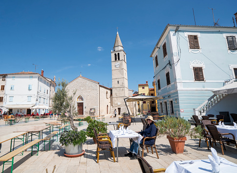 View of mature woman enjoying central square of small coast town Fazana on Istria coast while sitting on chair and waiting for lunch, Croatia. In background is blue sky, church in center and restaurants on both sides as well as some market tables. Fazana is starting point of boat tours to Brioni island where use to be residence of former Yugoslav president Tito.