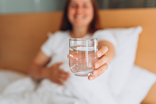 Pregnant woman with glass of water sitting on bed at home. Millennial Caucasian woman wake up in cozy white bed in modern apartment in the morning. Concept of healthy lifestyle