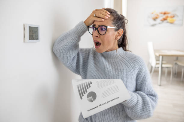 woman cannot believe her own eyes the sum on her energy bill looking shocked at the thermostat on the wall next to her. - power saving imagens e fotografias de stock