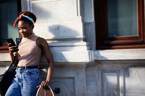 Portrait of happy african-american woman using the phone. Young stylish woman with skateboard outdoors