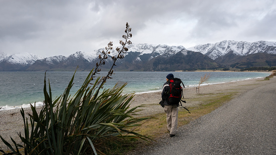 Man walking on the shore of lake Hawea in strong wind. Snow-capped mountains in the background, South Island.