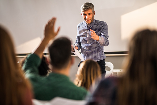 Smiling mid adult professor aiming at one student who wants to ask a question on a class at lecture hall.