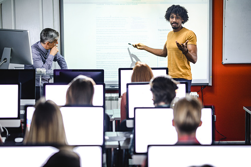 Young black student talking to his classmates in front of projection screen at computer class.
