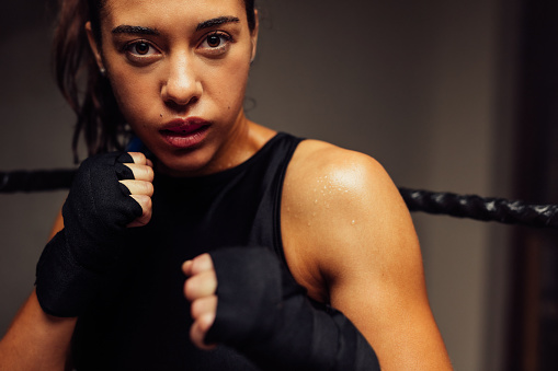 Female martial artist looking at the camera while standing in fighting position inside a boxing ring. Female boxer training in a boxing gym.