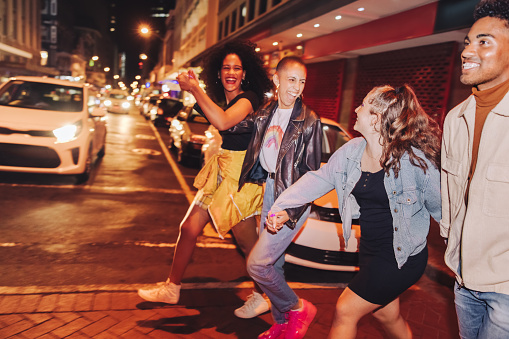 Group of multicultural friends crossing a road together at night. Happy friends smiling cheerfully while going out together in the city. Vibrant young people having a good time on the weekend.