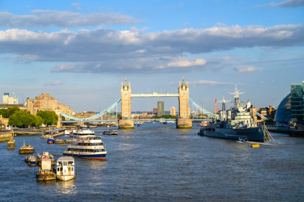 Wide angle view of boat traffic on water, moored warship, and landmark 19th-century bridge crossing near Tower of London on sunny summer day.