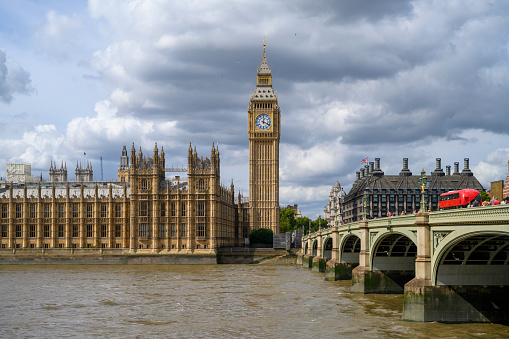 View across River Thames of Palace of Westminster, Elizabeth Tower with landmark clock face, and commuters crossing green arch bridge under dramatic summer sky.