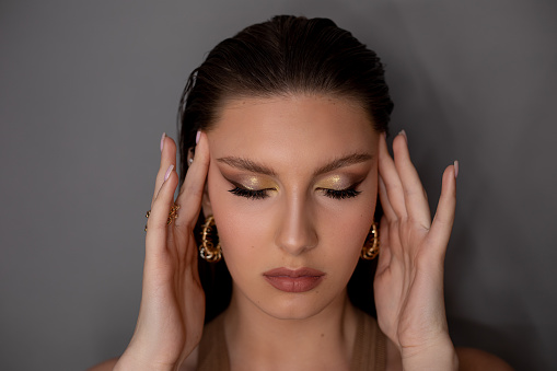 Portrait of young perfect woman with dark hair, bright golden make-up, pink lips, wearing round golden earrings with closed eyes, touching temples on grey background. Studio shot. Beauty, cosmetics.