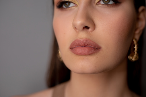 Close-up of young mysterious green-eyed woman with long dark hair, bright golden make-up, pink lips, wearing big round golden earrings on grey background. Studio shot, soft focus. Beauty, cosmetics.