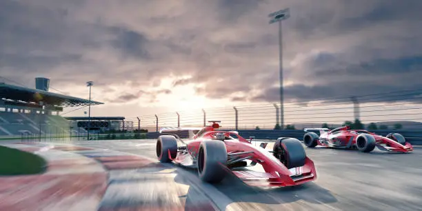 Two generic racing cars moving at high speeds, driving around a corner of a racetrack at sunrise/sunset under a cloudy sky, with empty spectator stands in the background. With motion blur to background and lens flare.