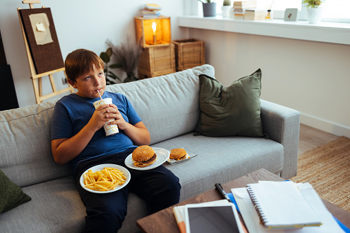 Overweight teenage boy sitting on a sofa and drinking juice using straw.