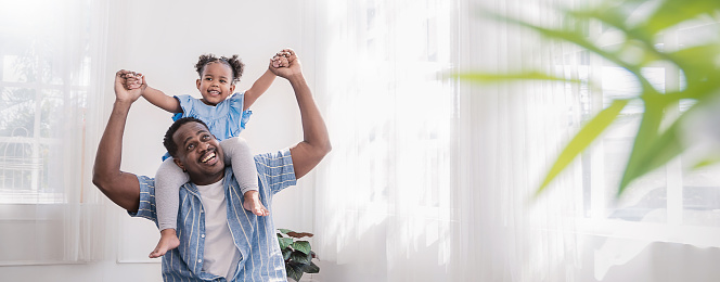 Happy black African American father daughter playing at home living room. Afro man carry piggyback little toddler girl. Cheerful family bonding together fatherâs day concept banner with copy space