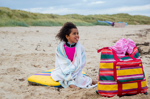 A shot of a young girl at a beach in Beadnell, Northumberland. She is sitting on a bodyboard wearing a wetsuit and she has a towel wrapped around her. She is sticking her tongue out.