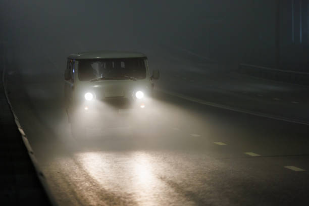 Two cars drive towards the camera with headlights illuminating a wet road, on a foggy and snowy mountain.