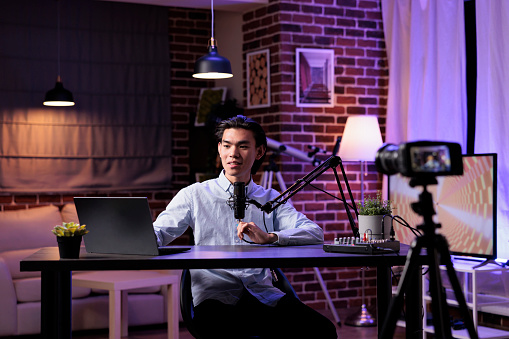 Young adult livestreaming talk show on social media channel, using sound equipment and laptop at studio station. Filming online video blog with podcast production, recording vlog content.