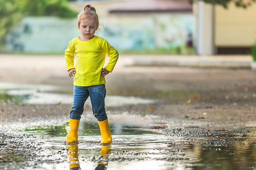 a little girl in yellow clothes and rubber boots cheerfully runs through the puddles after the rain in the warm season on a walk