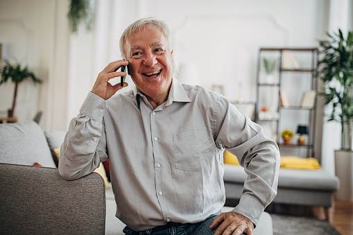 Senior man sitting on sofa alone in his living room, he is talking on mobile phone.