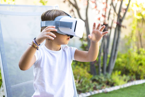 Little girl with virtual reality glasses, looking to the right, trying to touch something in a virtual way, sitting on a sun lounger in the garden of her house. Metaverse, VR, game, digital concept. stock photo