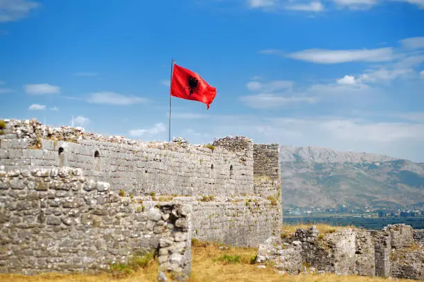 Photo of Red Albanian flag with double-headed black eagle waving over wall of Fortress Rozafa near Shkodra city. Fluttering banner with symbol of Albania on background of sky. Travel and tourism.