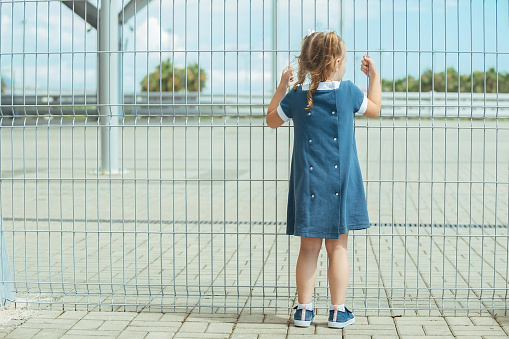 a little preschooler girl in a blue dress stands near a barrier from a high mesh fence and holds on to it with her hands