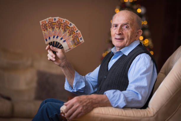 Happy old man holding a fan of money Cheerful grandfather sitting in a chair with a lot of money in his hand euro bill in a christmas ornament stock pictures, royalty-free photos & images