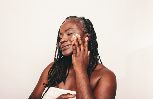 Mature woman with dreadlocks applying moisturizing cream on her face. Confident dark-skinned woman taking care of her flawless melanated skin. Mature black woman ageing gracefully.