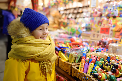 Cute little boy having wonderful time on Xmas market. Child choosing souvenirs, presents and gifts for family and friends . A lot colorful and multicolor wooden toys at Christmas fair in the big city.