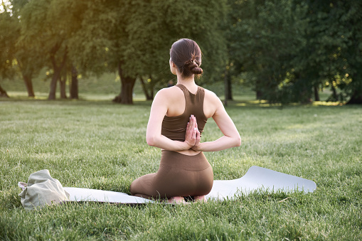 A young positive woman in a gymnastic suit practices yoga and meditates while sitting on a mat. Rear view. Active and healthy lifestyle concept.
