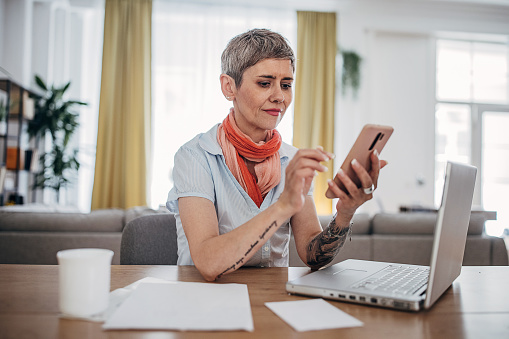 One woman, mature female sitting at the desk and using smart phone in her home office