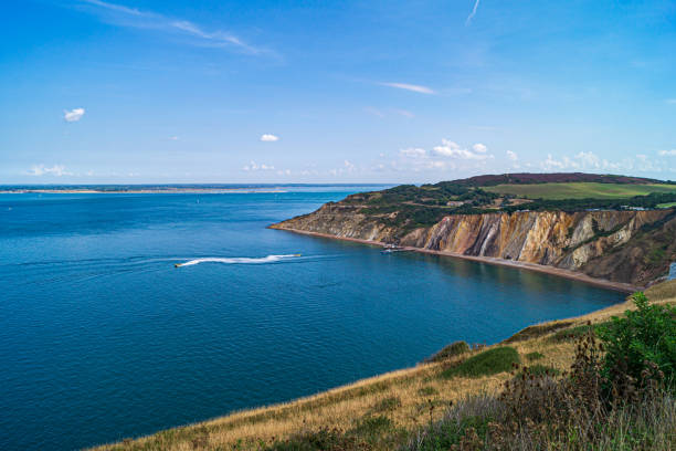 A view of Alum Bay, Isle of Wight taken from the cliffs above. The seas are calm and a small boat can be seen leaving the jetty. The cliffs are reflected in the calm sea.